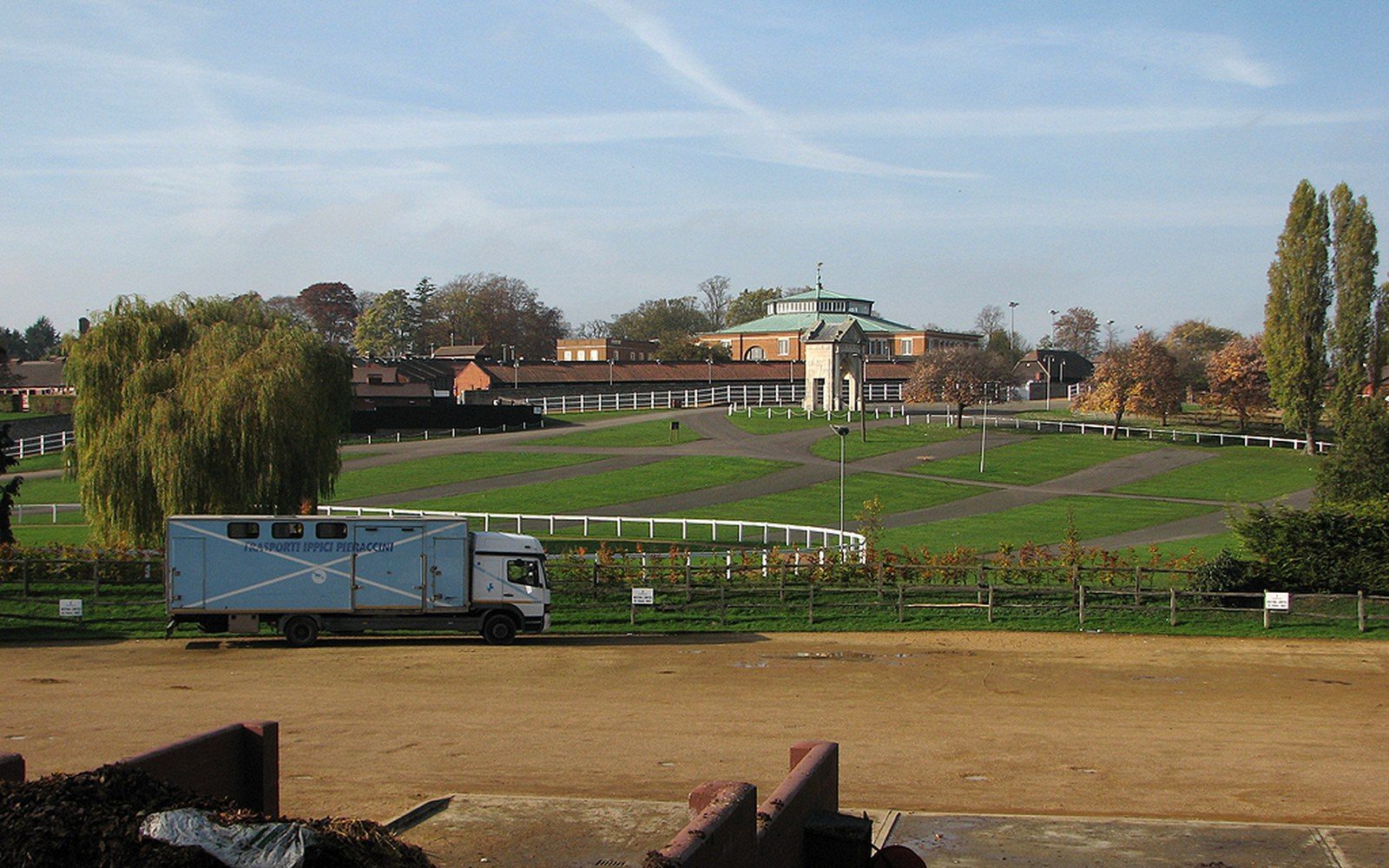 A horse transport lorry parked beside white-railed paddocks with a domed pavilion in the background